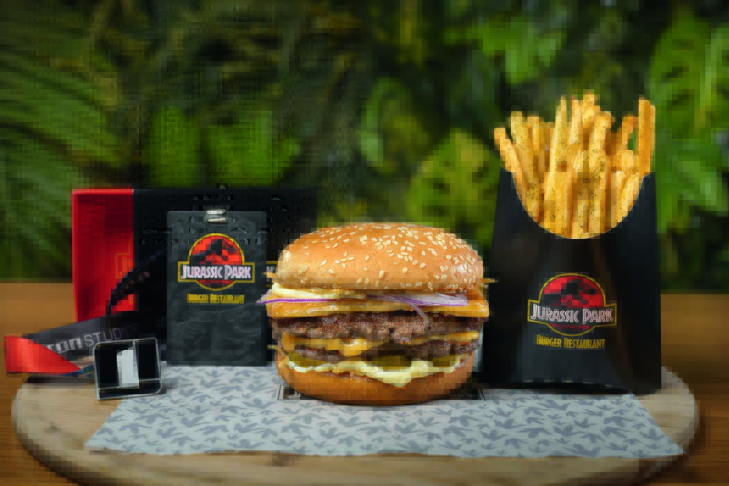 hambúrguer com dois discos de carne prensados, três fatias de queijos da casa, cebola-roxa, maionese e picles de pepino, além de batatas fritas, um pin e uma credencial que remete à embalagem da fita VHS do longa Jurassic Park