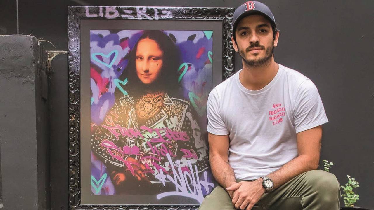 Homem branco de boné cinza e camiseta branca sentado em cadeira com Monalisa colorida ao fundo.