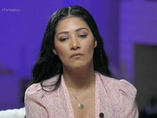 Simaria aparece com semblante sério em estúdio de entrevista, com fundo azul, usando camisa rosa