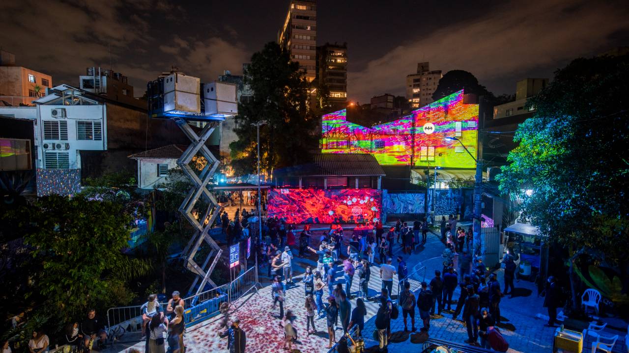 Beco do Batman envolto em projeções a laser de imagens coloridas. Pessoas olham e tiram fotos. O espaço é visto de cima, durante a noite