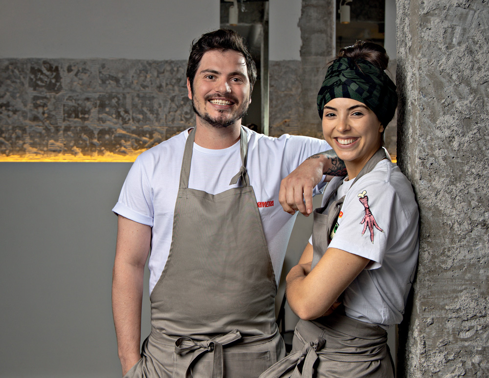 2022_08_28_Comer_e_Beber_Gabriel_Júlia_Wanezza_Soares-6516-Edit.psd Imagem mostra homem e mulher sorrindo, ambos com avental bege sobre camiseta branca