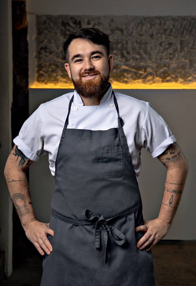2022_08_28_Comer_e_Beber_Raphael_Vieira_Wanezza_Soares-6663-Edit.psd Imagem mostra homem barbudo de camiseta branca e avental cinza, sorrindo.