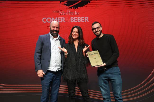 Comer_&_Beber Mulher vestida de preto entre dois homens, um deles com uma placa