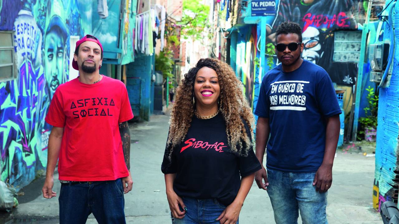 Imagem mostra dois homens e uma mulher em viela de uma favela