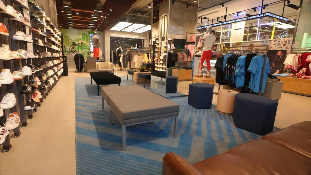 Adidas – Loja-conceito no Top Center (DIVULGAÇÃO) (1) Sofás e pufes em loja com tapete azul e cinza. Manequins com roupas esportivas ao fundo.