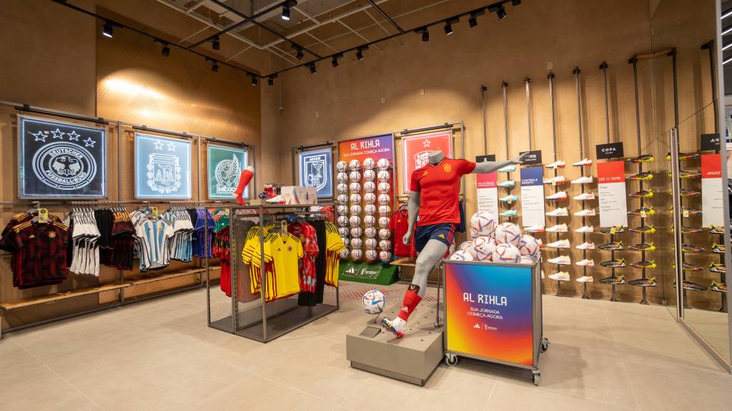 Adidas – Loja-conceito no Top Center (DIVULGAÇÃO) (5) Foto exibe araras com camisas da seleção brasileira e manequim de jogador de futebol com uniforme laranja e azul e bola no pé.