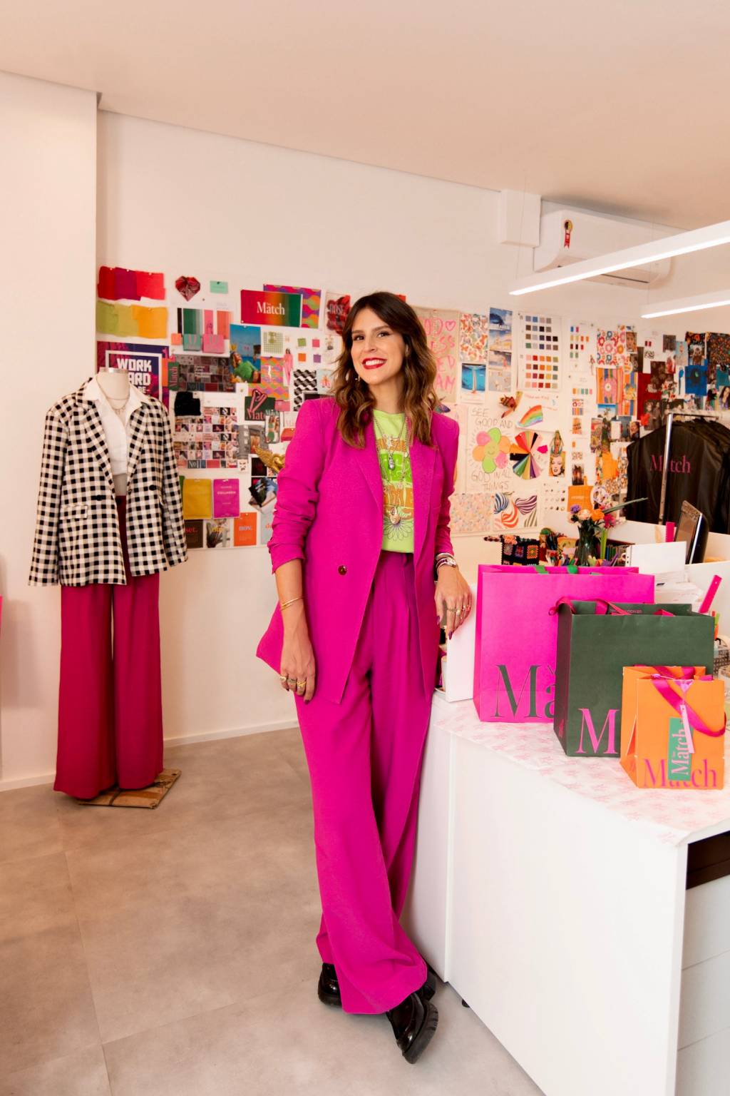 Ciccy Halpern (Juliana de Souza) (3) Ciccy Halpern posa sorrindo com blazer rosa pink e sacolas em primeiro plano. Ao fundo, mural repleto de papéis coloridos.