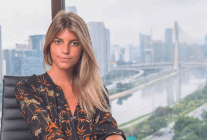 Imagem mostra mulher loira sentada em mesa. Ao fundo, uma janela mostrando a Ponte Estaiada, em São Paulo