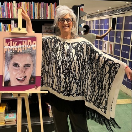 Kika Gama posa de braços abertos com roupa estampada preta e branca, cabelos brancos, óculos de grau. Está ao lado de um pôster que estampa seu livro.