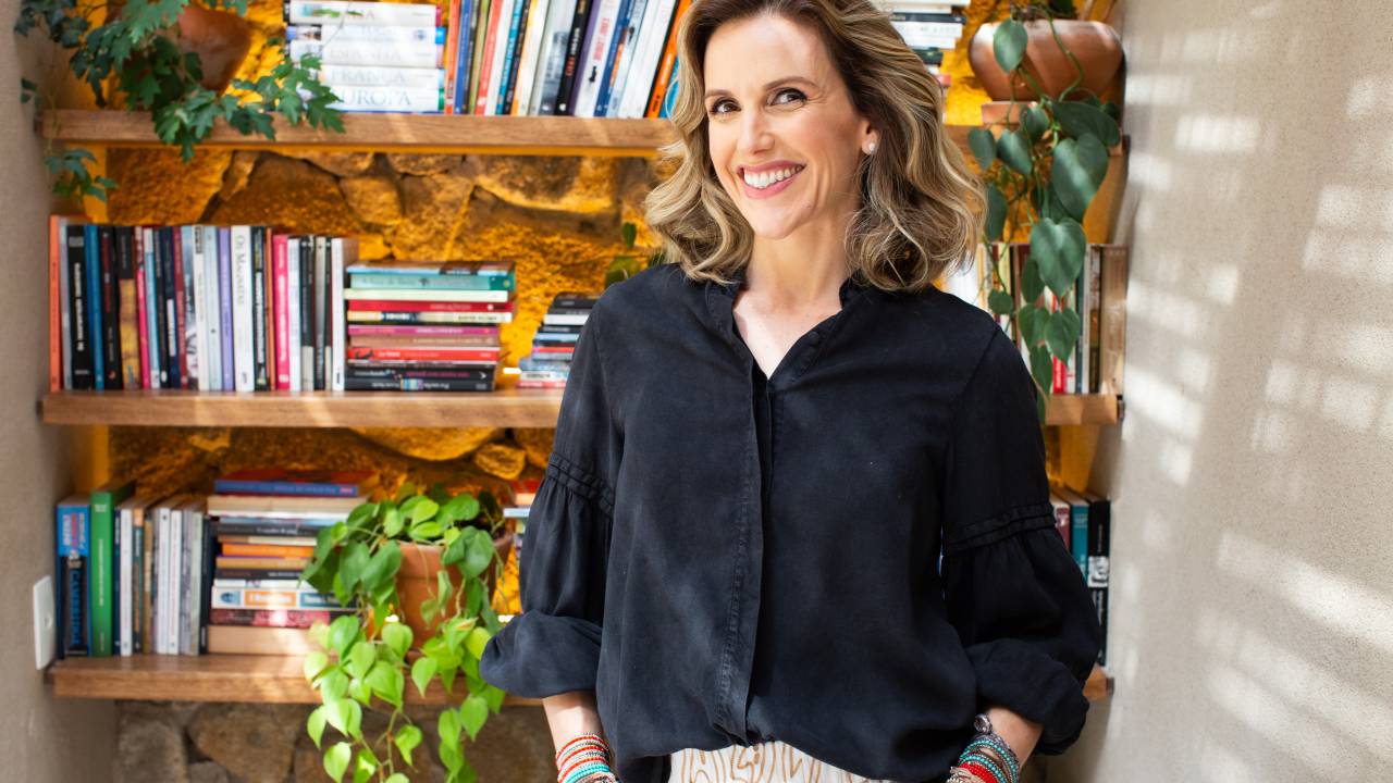 Mariana Ferrão sorri em local com prateleiras de livros e plantas. Veste camisa preta.