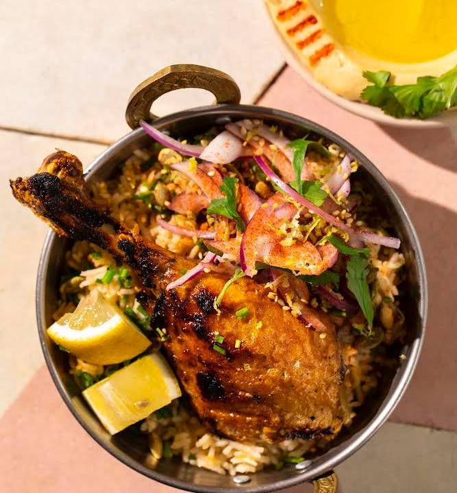 Panelinha de ferro sobre mesa rosa contendo o kabsa, que leva arroz basmati, galeto assado e caldo com especiarias