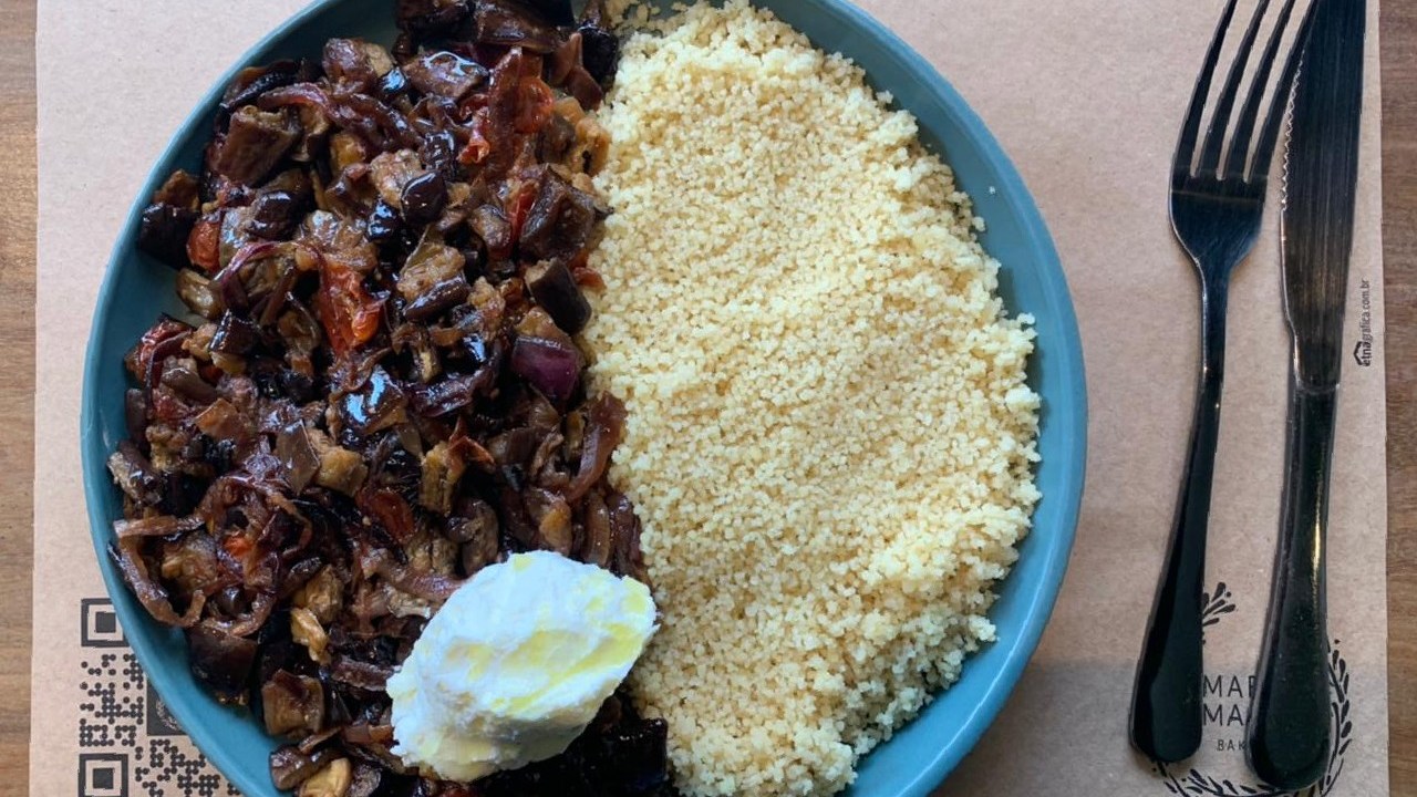 Prato azul sobre jogo americano pardo com garfo e faca do lado direito, dentro do qual há caponata de berinjela, cebola roxa e tomate, cuscuz marroquino e coalhada seca