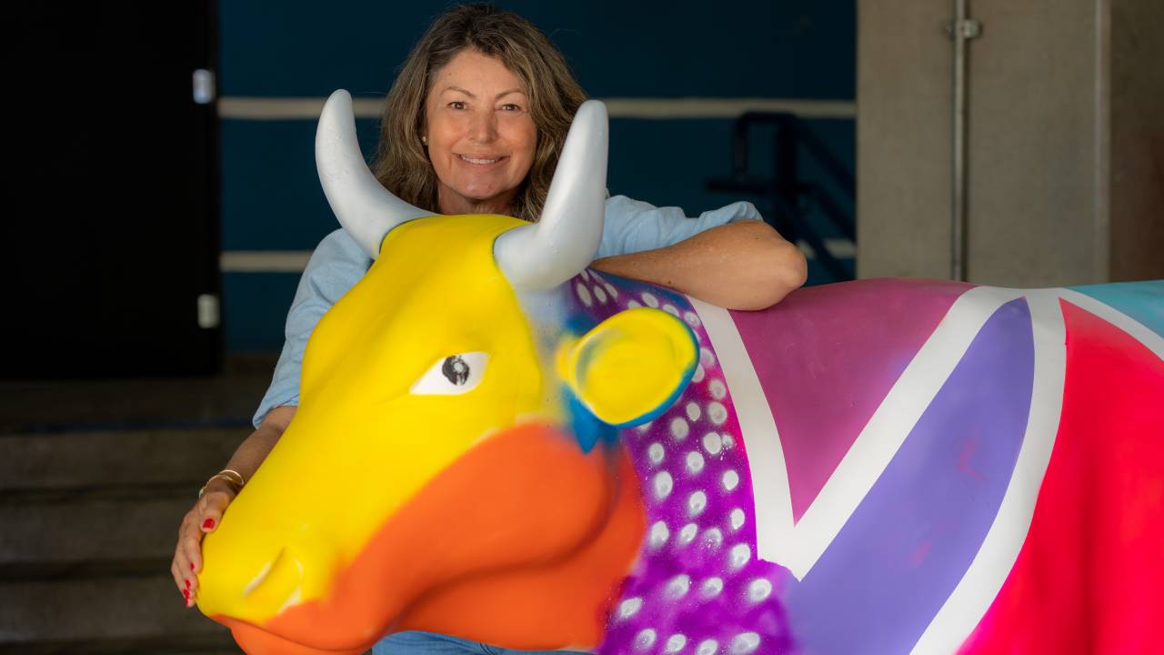 Catherine Duvignau posa atrás de escultura de vaca pintada nas cores amarelo, laranja, rosa, azul e roxo com pontinhas brancas.