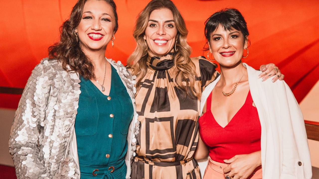 Três mulheres sorriem lado a lado. A primeira da esquerda tem cabelos longos escuros, veste blusa branca e conjunto verde. A do meio tem cabelos longos com luzes e veste conjunto estampado nas cores marrom e bege. A terceira veste blusa decotada vermelha com calça bege e mantinha branca nos ombros. Tem cabelos curtos escuros.
