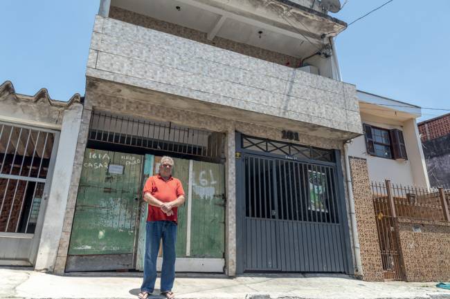 Foto de Adolfo, usando camiseta vermelha e calça jeans, em frente a sua casa