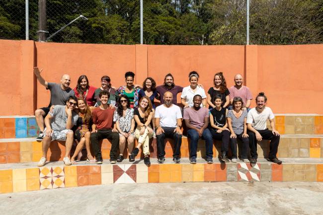 Professores sentados em arquibancada a céu aberto em 2 fileiras, cerca de 20 professores