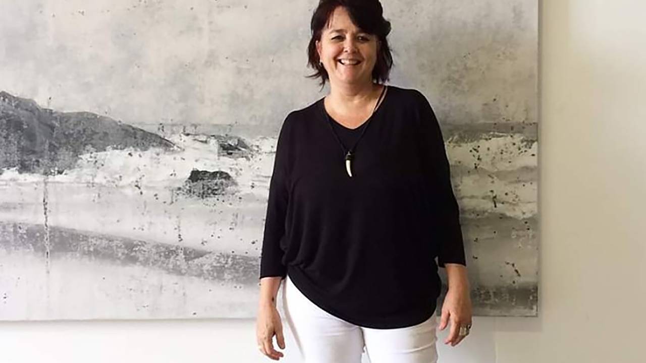 Mulher branca de cabelos curtos castanhos posa de pé em frente a quadro preto e branco de paisagem de praia. Veste calça branca e blusa preta de manga longa.