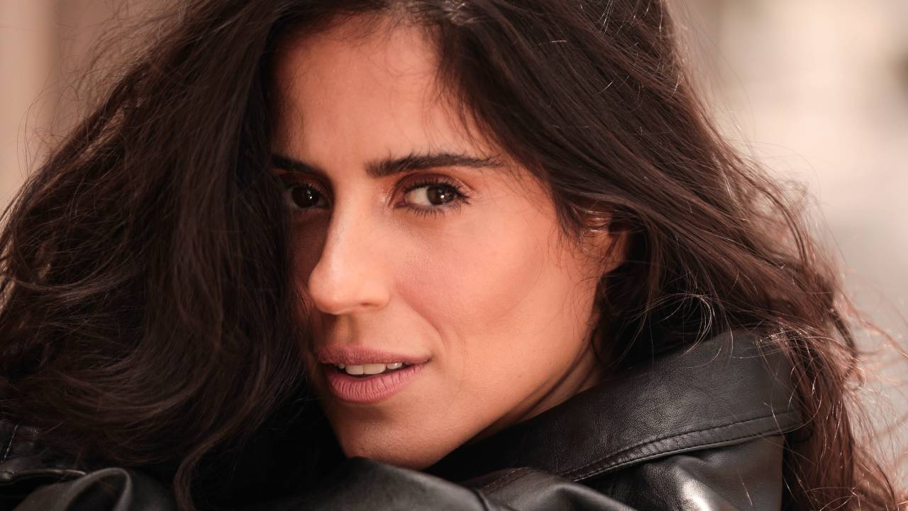Camilla Camargo posa com o rosto de lado em um meio sorriso. Os cabelos longos são castanho escuro e a jaqueta é de couro.