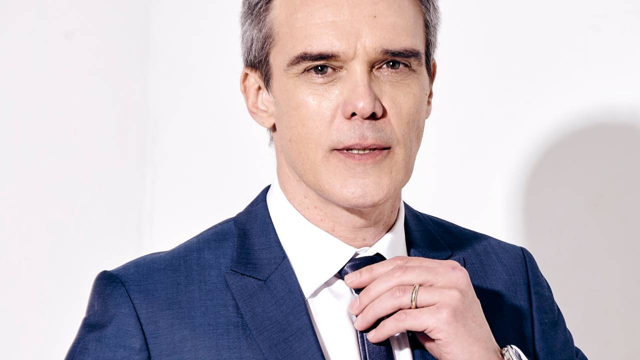 Homem grisalho posa segurando lapela do blazer azul com camisa branca e gravata escura.