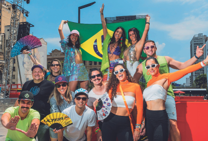 Imagem mostra grupo de pessoas vestindo roupas de carnaval, alegres, e segurando bandeiras e cartazes de festival de música. Ao fundo, um céu azul.