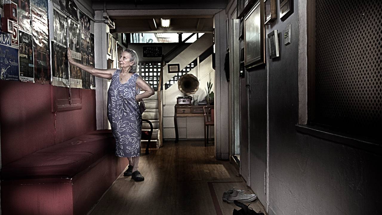 Imagem mostra mulher idosa apoiada em parede, usando vestido