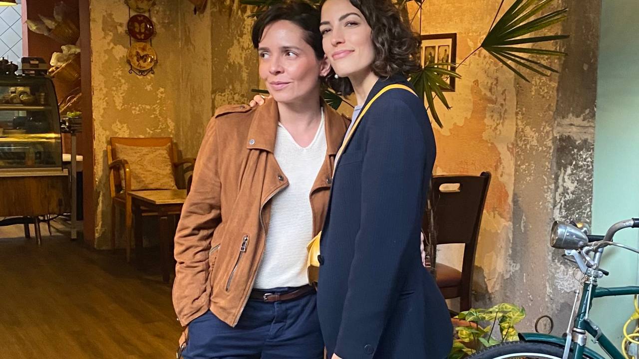 Nadia Bochi e Sílvia Henz posam de pé abraçadas, ambas com calças escuras, Silvia à direita com blazer preto e Nadia com camisa branca e jaqueta marrom. Ao lado aparece uma bicicleta.