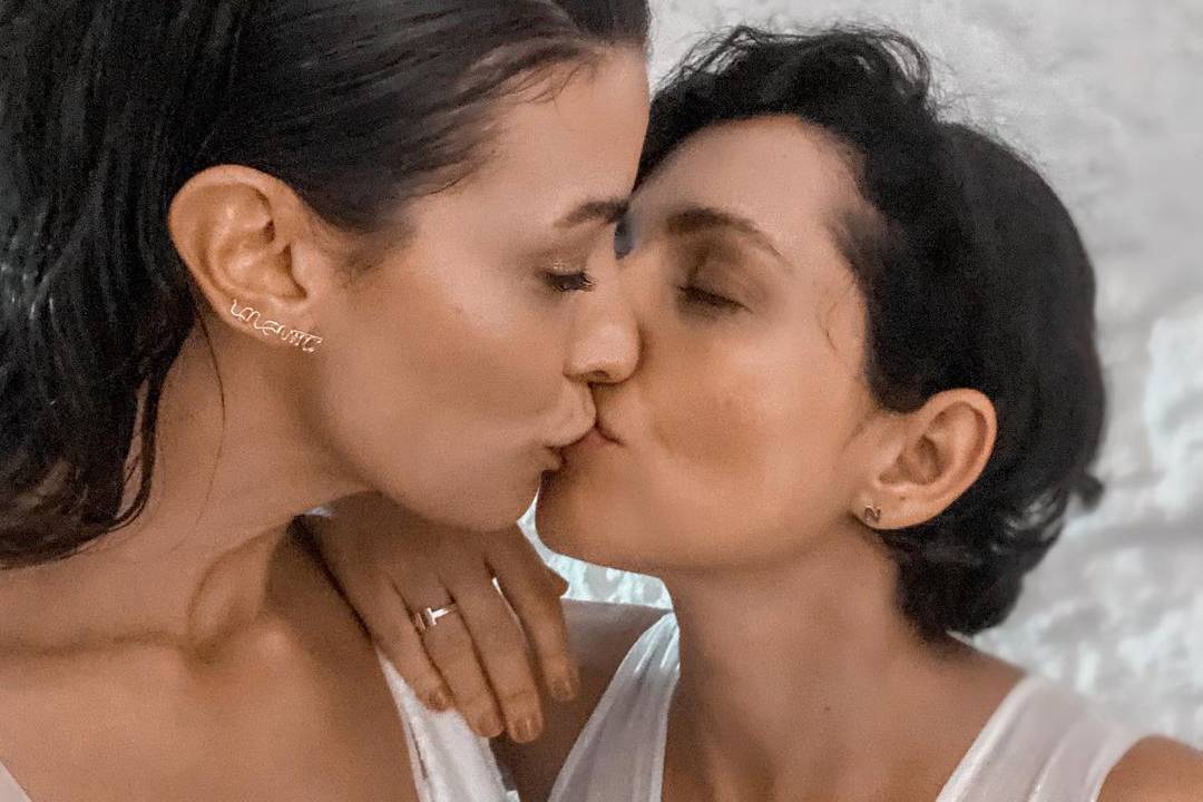 Duas mulheres de cabelos curtos e pretos aparecem dando selinho de olhos fechados. Ambas usam regatas brancas.