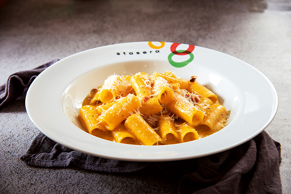 Carbonara feito com rigatoni em prato fundo onde se lê Stasera