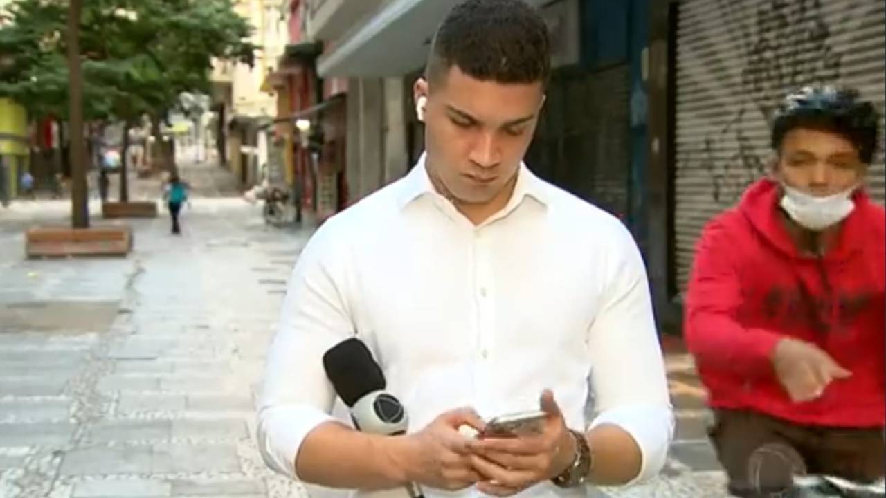 Imagem mostra homem de camisa branca, olhando o celular, e homem em bicicleta atrás