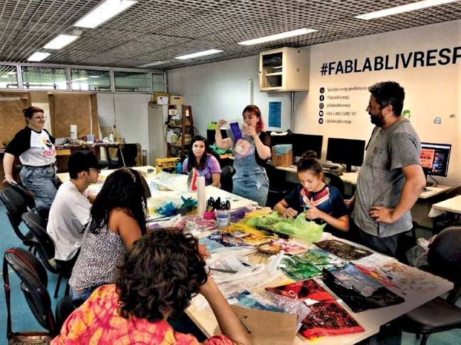 Adereços Plastico Preciosos – Fab Lab Penha – 2020 (2).jpg Adultos e jovens sentados ao redor de uma mesa acompanham explicação que uma pessoa dá em pé