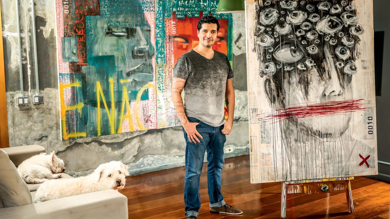 Imagem mostra homem de camiseta cinza e branca e calça jeans, em pé, com as mãos nos bolsos. No espaço, cachorros em um sofá e três grandes telas de pinturas ao redor do homem