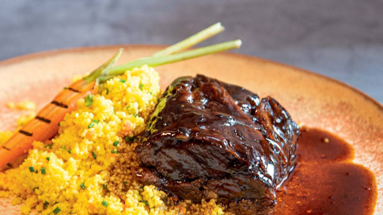 Prato de cerâmica sobre mesa azul, com paleta de cordeiro, cuscuz marroquino temperado e uma minicenoura com marcas de grelha