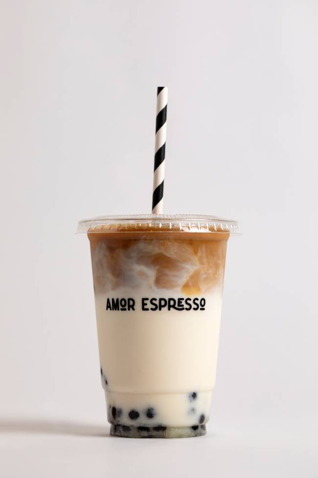 Copo transparente onde se lê Amor Espresso em letras pretas, com parte clara que parece leite e parte escura que parece café. Ao fundo, bolinhas que são bobbas ou bolinhas de tapioca. Canudo é listrado de branco e preto