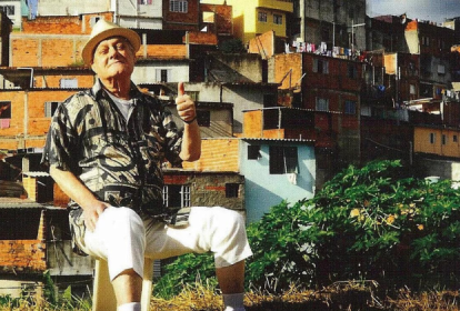 Imagem mostra homem de chapéu, sentado em cadeira em frente a comunidade