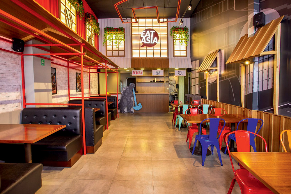 Ambiente interno do restaurante com coberturas remetendo a estabelecimentos e casinhas, paineis com referências ao Godzilla, mesas e cadeiras