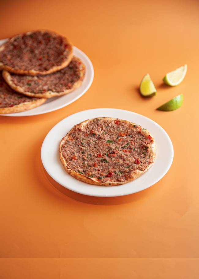 Esfirras de carne aberta Yamadjian pilha de esfirras abertas de carne sobre pratos em cima de mesa laranja, com limão cortado em cunhas ao lado, sobre a mesa