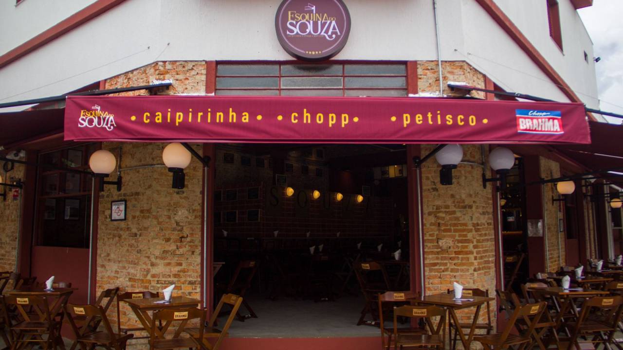 Entrada de boteco, com toldo vinho onde se lê caipirinha, chopp e petisco, situada numa esquina. Na foto aparecem as laterais da entrada em tijolinhos à vista e uma mesinha de cada lado da porta