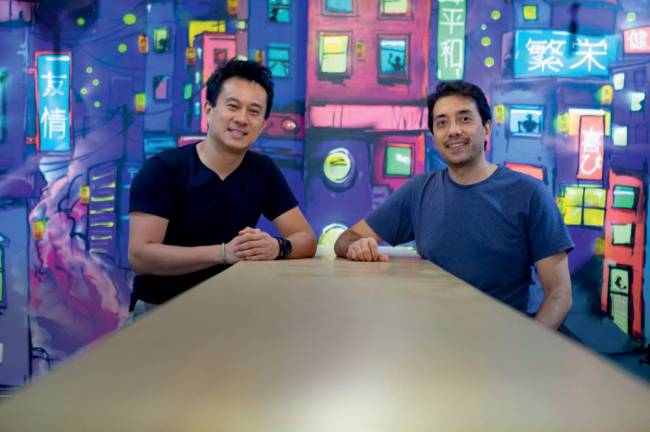 Ricardo Fan e Henry Nakaya Eat Asia Ricardo Fan e Henry Nakaya sentados à mesa em frente a parede desenhada em 2D