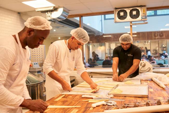 Cozinha Le Jazz Boulangerie Pessoas trabalham em cozinha sobre mesa de madeira, usam toucas e aventais