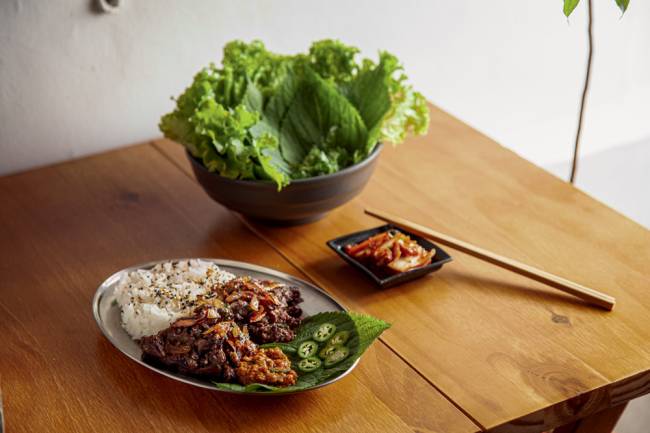Bugolgi com seus acompanhamentos Wooza Prato com bulgogi e arroz e tigela de metal com folhas para fazer as trouxinhas