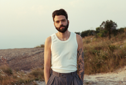 Imagem mostra homem de regata branca e barba, em paisagem de grama