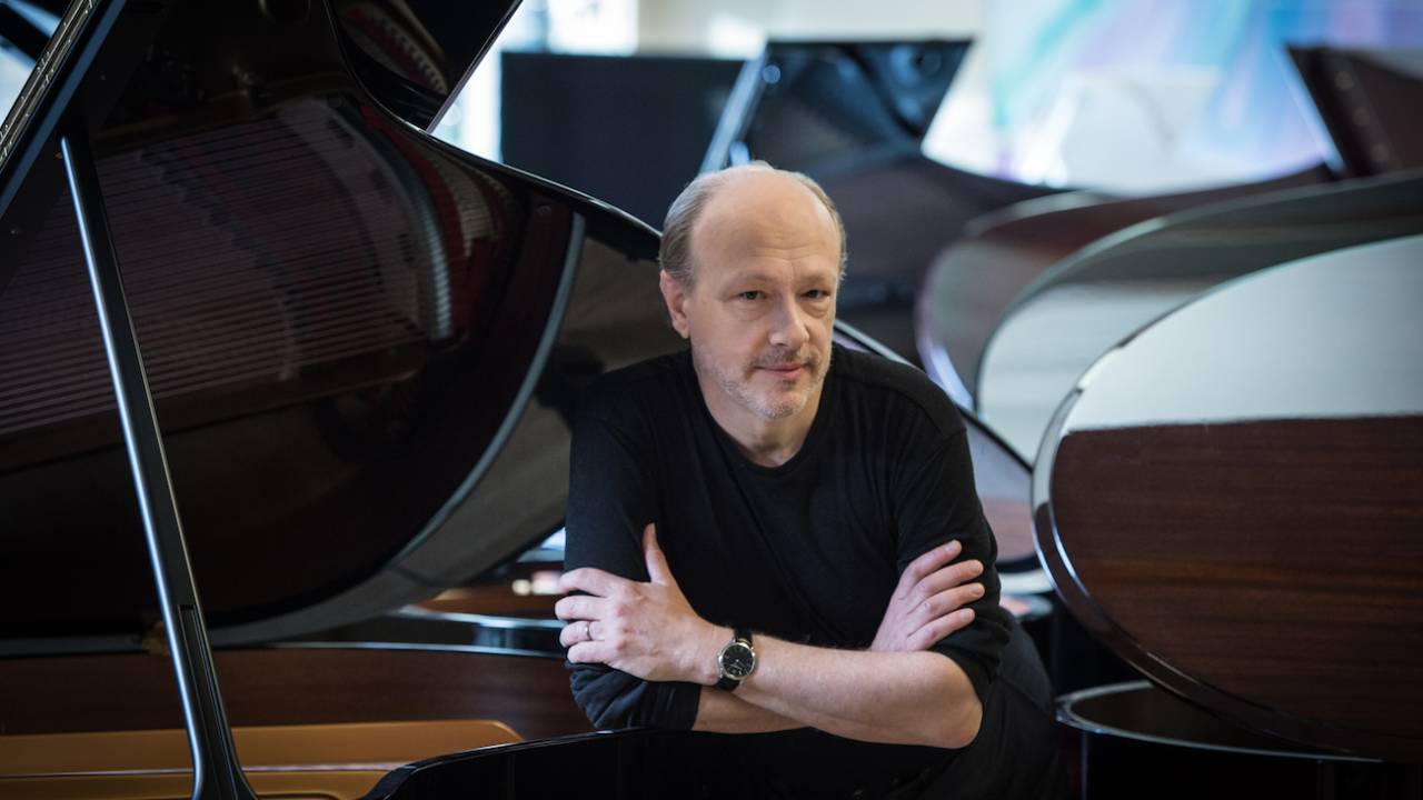 Imagem mostra homem careca apoiado em piano, vestindo roupa preta