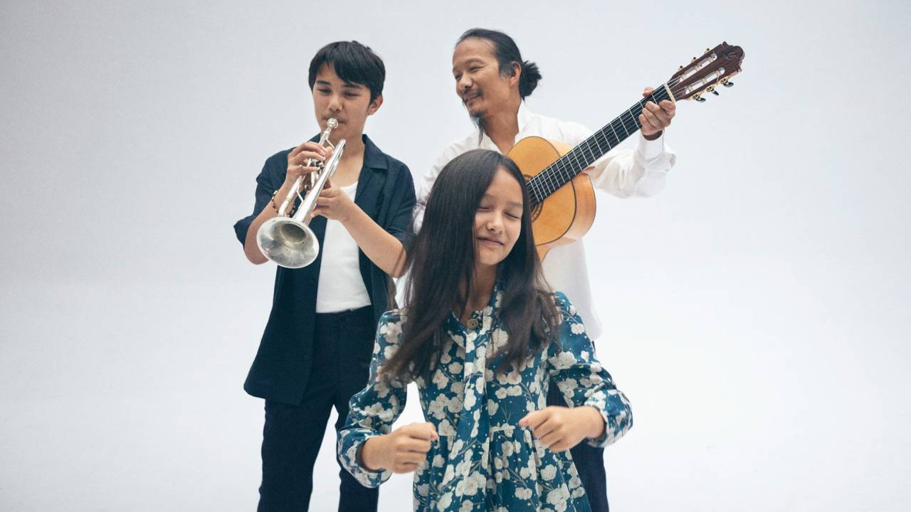 Imagem mostra três pessoas sobre fundo branco, em pé, tocando instrumentos