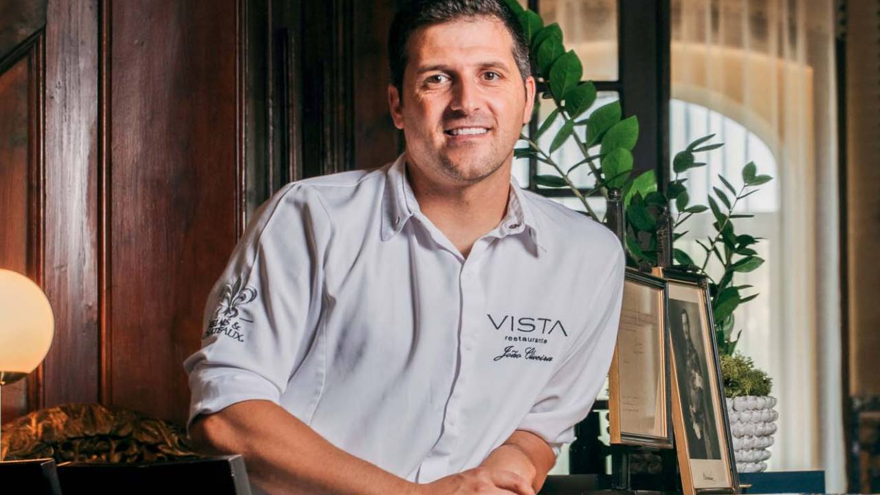 Homem sentado em cadeira escura vestindo camisa cinza sorri para a câmera