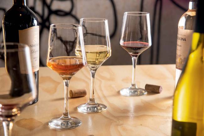 Le Bon Vin 012.jpg Três taças contendo vinhos branco, rosé e tinto sobre mesa de madeira clara