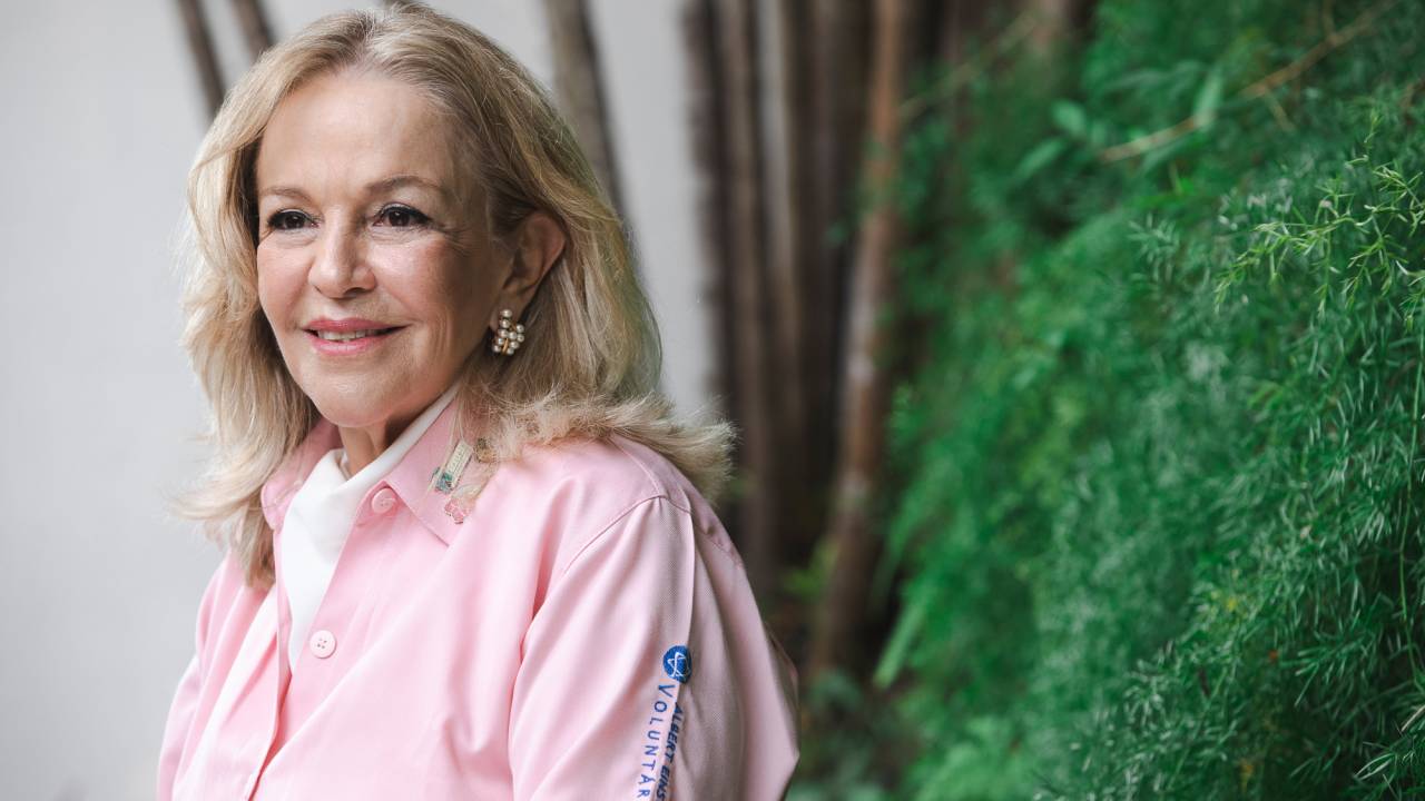 Senhora branca e loira sorri olhando para a esquerda. Veste blusa rosa claro.