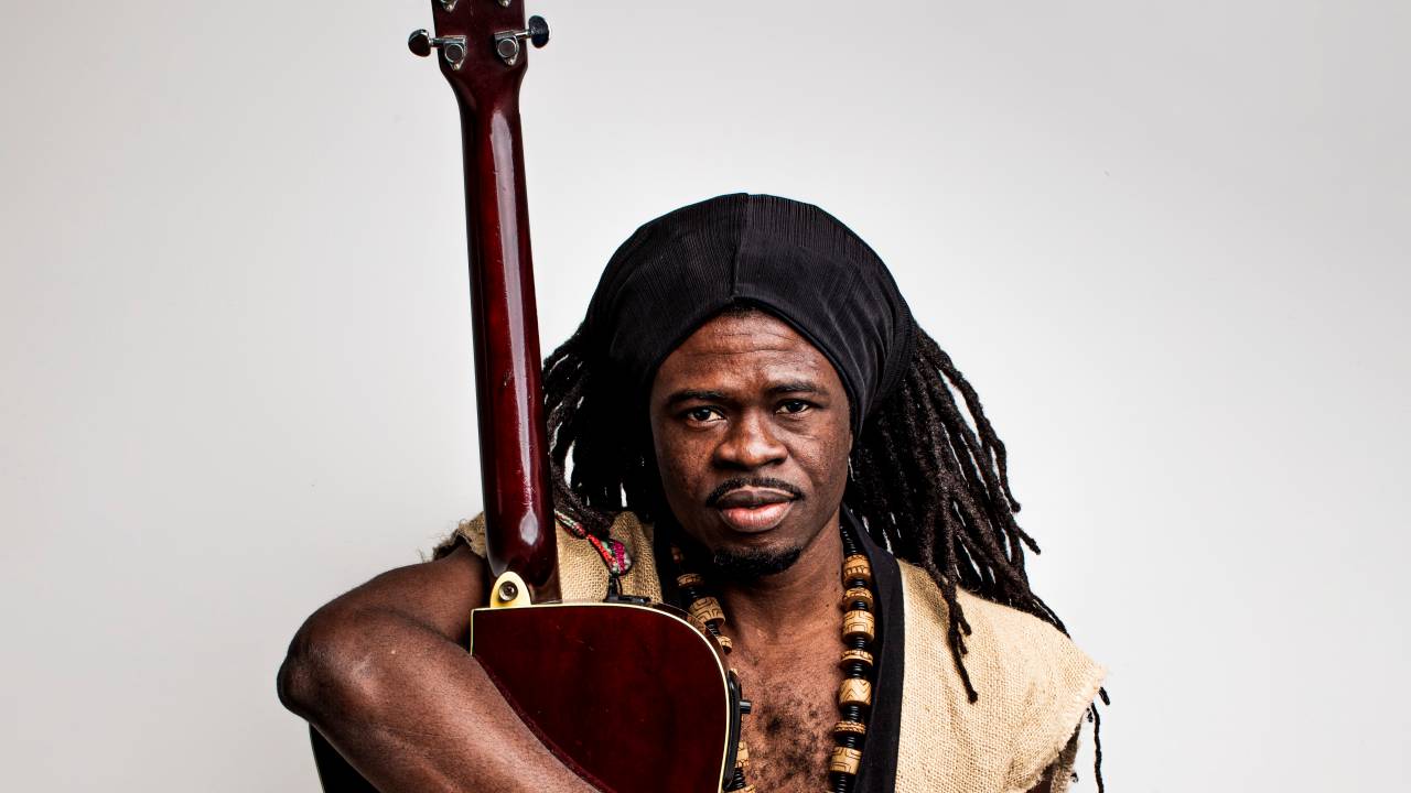 Cantor e compositor Yannick Delass posa abraçado com violão de pé. Tem dreads, pele negra e veste um colete cor de palha.