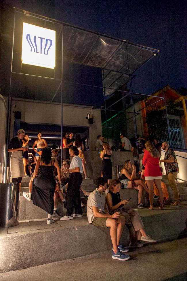 Arquibancada Bar Alto Arquibancada com pessoas em pé e sentada conversando, e letreiro do bar alto acima à direita