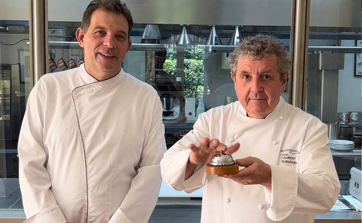 Dois chefs vestindo dolma olham para a câmera, um deles segura uma campainha de metal de balcão nas mãos