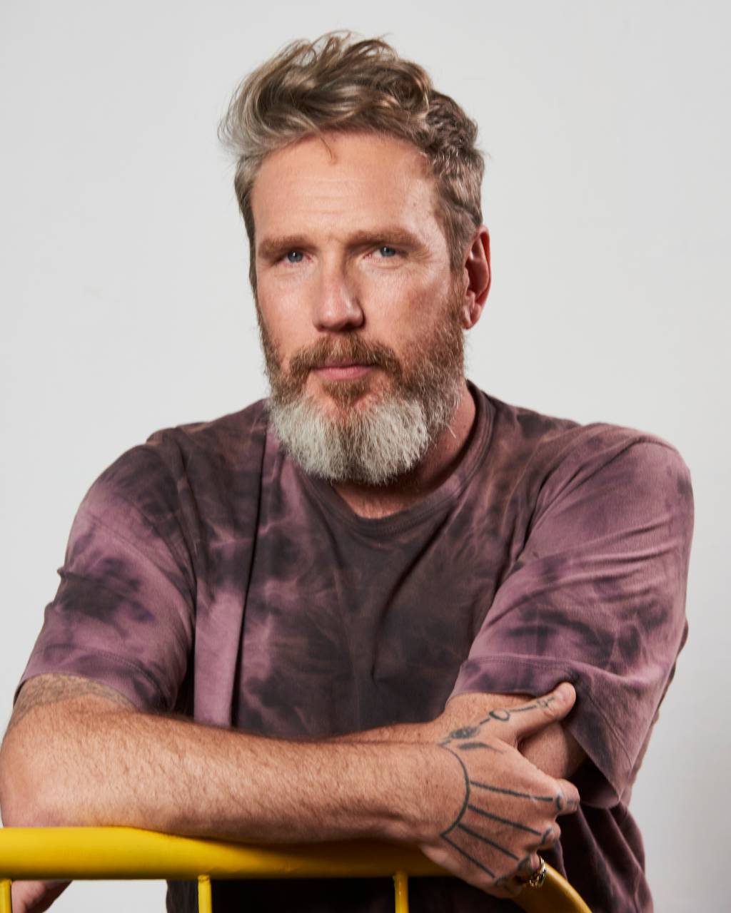 Alexandre Herchcovitch (Zé Takahash-Divulgação) Homem branco de barba e cabelo grisalhos encara a câmera sério com os braços levemente cruzados. Veste camiseta com tom manchado na cor roxa.